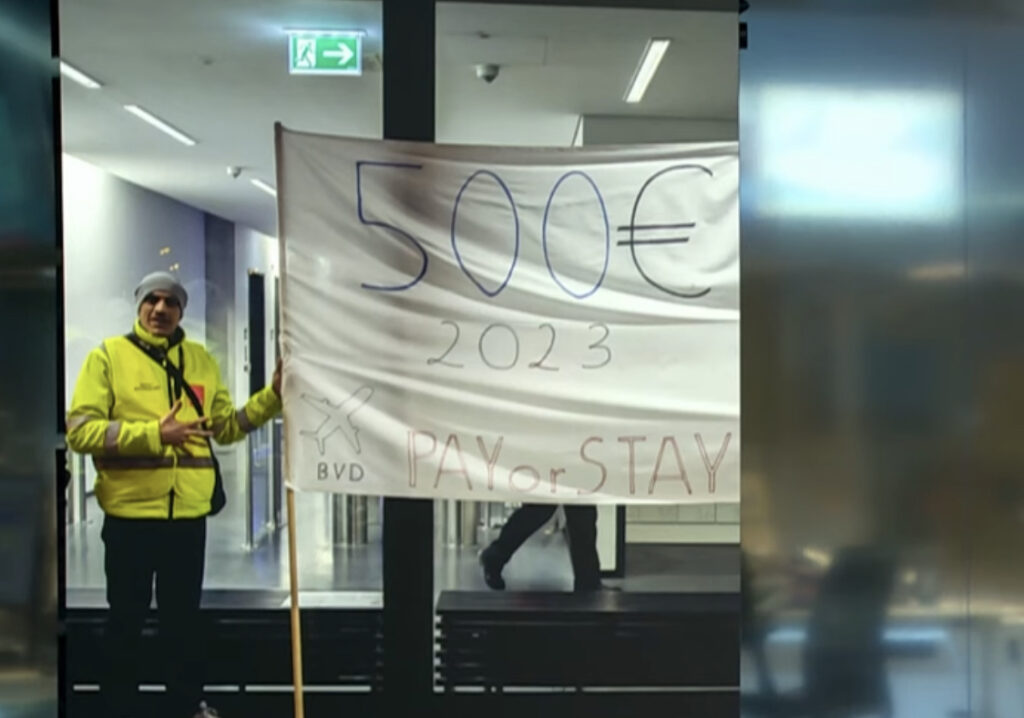 Eine Person in einer Warnweste hält ein selbstgemachtes Banner mit der Aufschrift „500€ 2023 PAY or STAY“ hoch