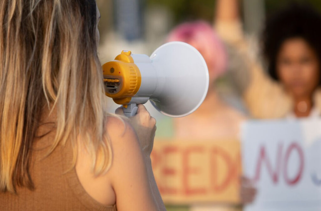 Person holding a megaphone at a protest, with activists and signs blurred in the background
