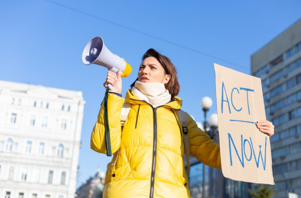 woman in yellow jacket with a megaphone holds a paper table with act now written on it